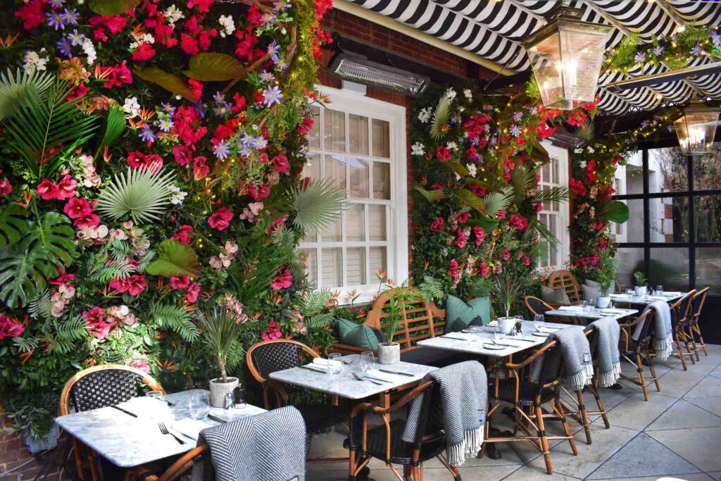 Adorable restaurant en plein air avec des murs fleuris et des couvertures sur les chaises en osier pour garder les clients au chaud, ainsi qu'un chauffage bromique.